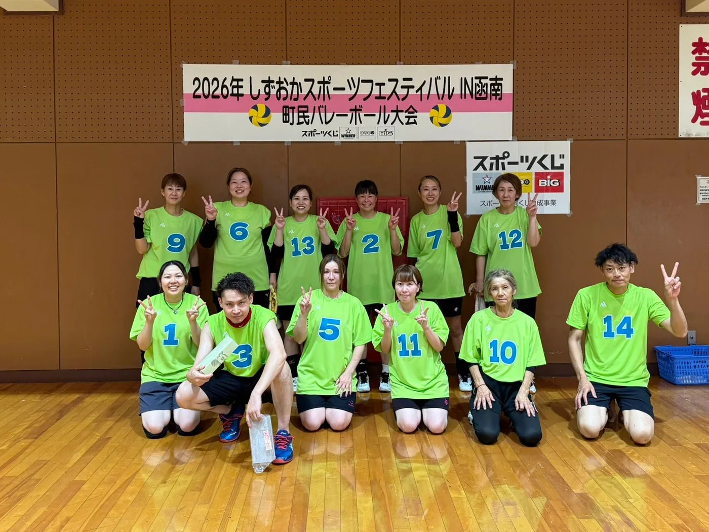 ライムグリーンに青い番号を入れたおそろいのTシャツで、体育館の大会横断幕の前に集合したバレーボールチームの選手たち