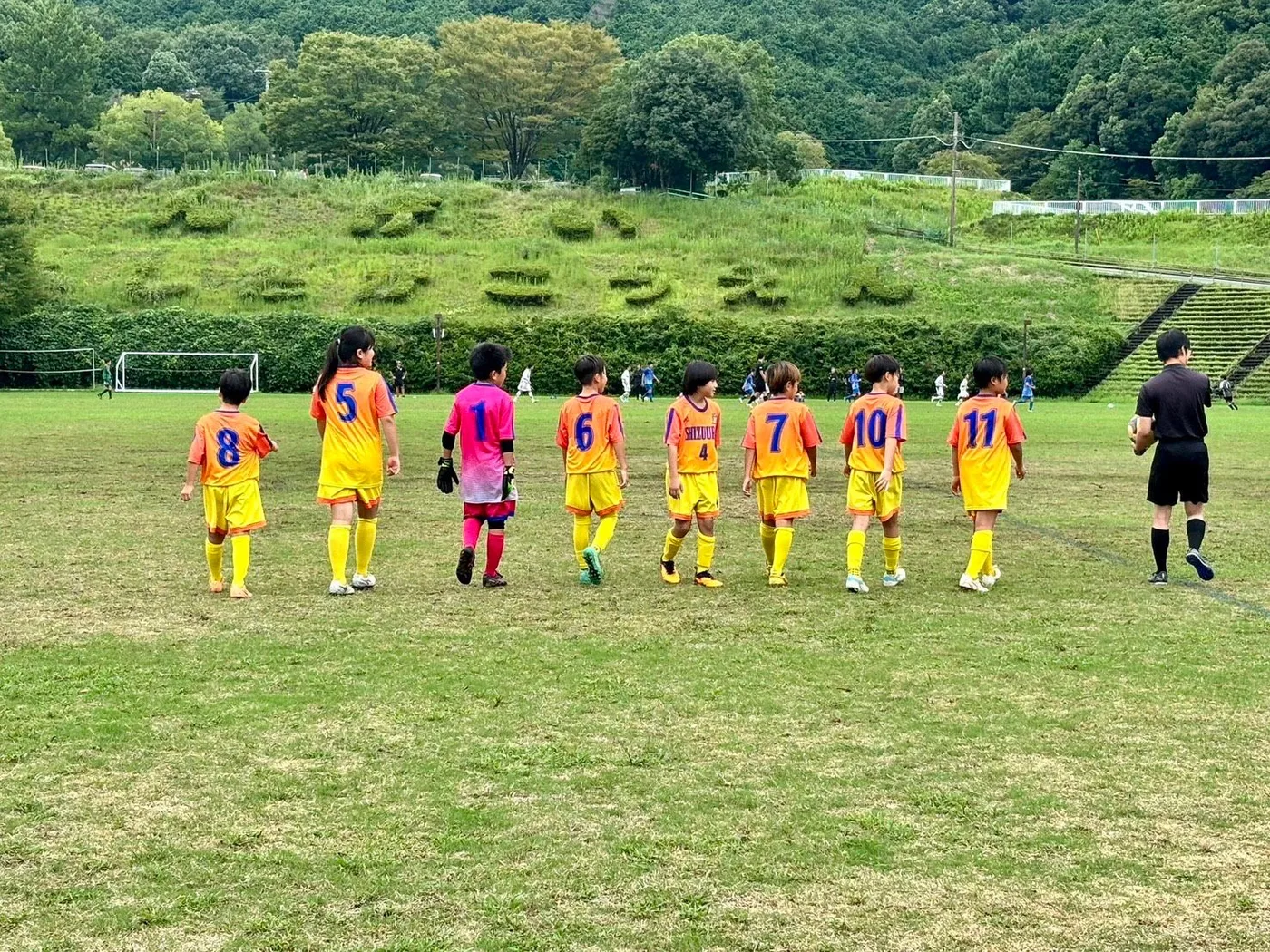 オレンジから黄色へグラデーションする昇華ユニフォームに黄色のパンツとソックスを合わせた静浦FCの選手たちが、ピンクのゴールキーパーと一緒に一列でピッチを歩いていく後ろ姿