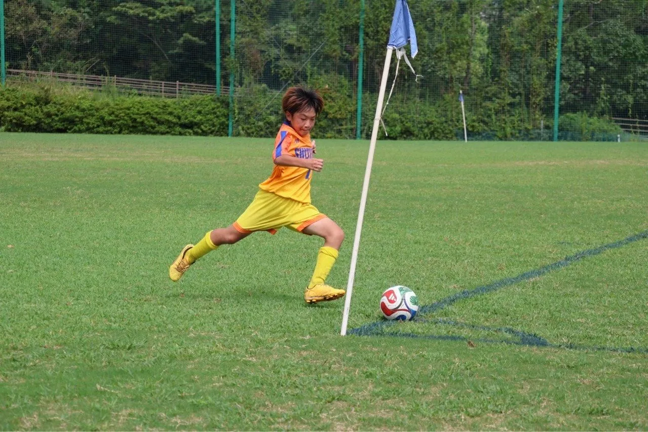 オレンジと黄色の昇華ユニフォームを着た静浦FCの選手が、コーナーフラッグの脇でボールを蹴ろうと踏み込む瞬間