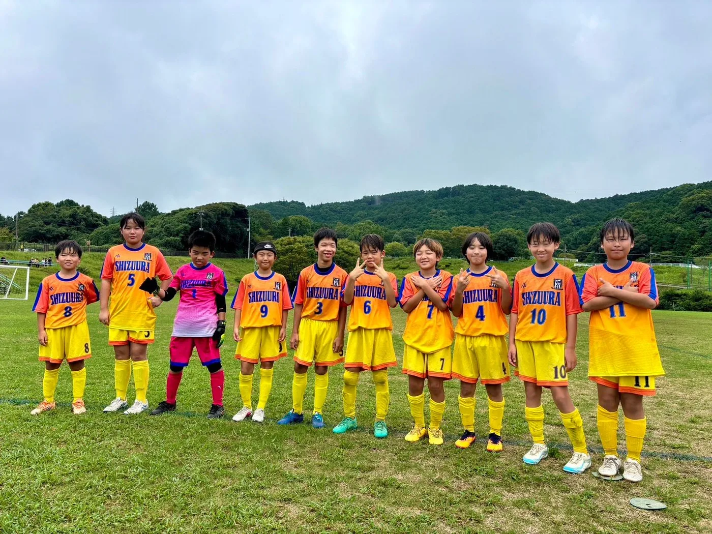 オレンジのグラデーションの昇華ユニフォームと黄色のパンツ・ソックスを着た静浦FCの選手10名が、ピンクのゴールキーパーと一緒にグラウンドで横一列に並んだ集合写真