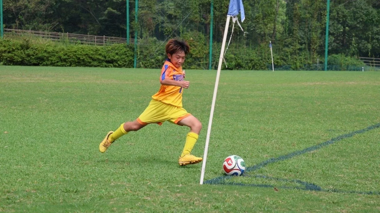 オレンジと黄色の昇華ユニフォームを着た静浦FCの選手が、コーナーフラッグの脇でボールを蹴ろうと踏み込む瞬間